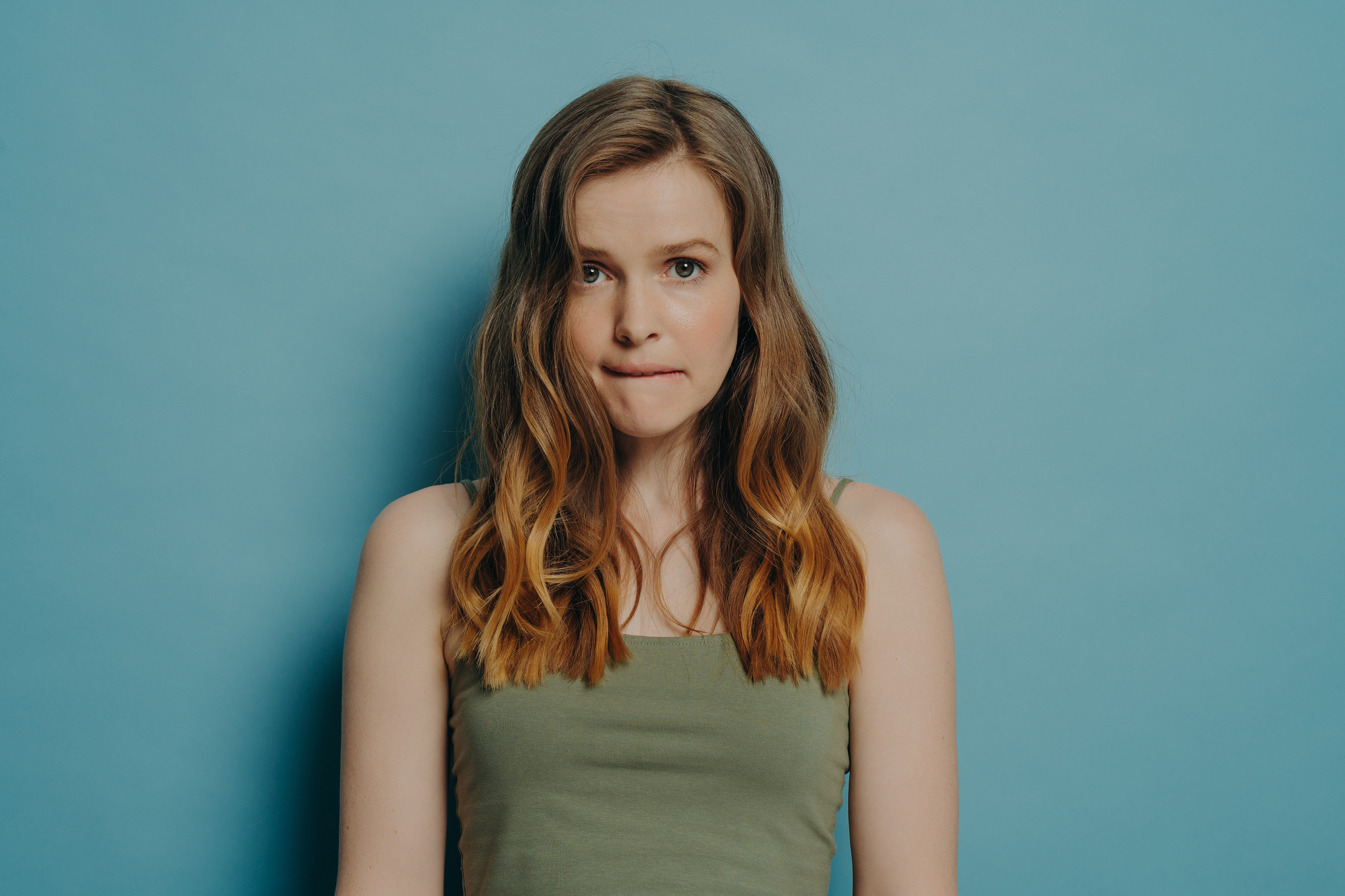 Confused worried young woman with wavy hair biting lip and feeling guilty, isolated on blue